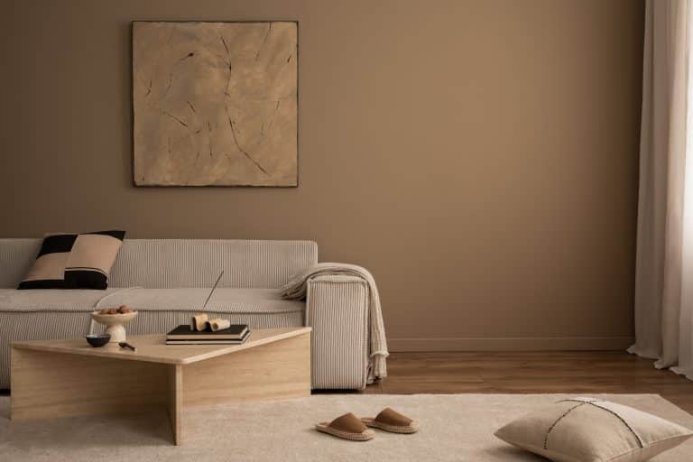 Salón cálido y acogedor con marco de póster, cuadro, sofá beige, mesa de centro de travertino, zapatillas, cuenco con frutos secos, manta a cuadros, cojín y accesorios personales.