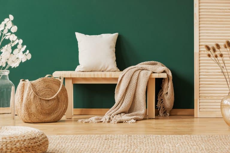 Wooden bench with beige carpet and pillow next to straw handbag, copy space on the empty green wall